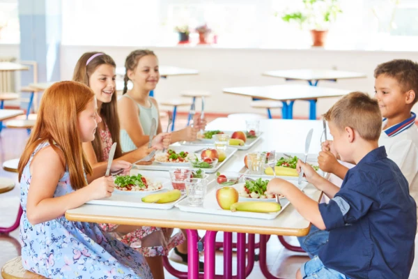 Happy kids having meal with cutlery while sitting together at table in school canteen