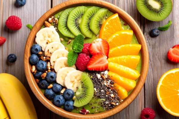 A top view of a vibrant green smoothie bowl with banana slices, blueberries, kiwi, strawberries, orange slices, raspberries, chia seeds, and granola, placed on rustic wooden background